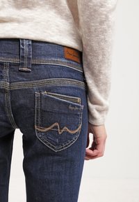 Vaqueros de mezclilla azul oscuro con un acabado texturizado, que presentan costuras en contraste y un parche de logo de cuero marrón en la cinturilla.