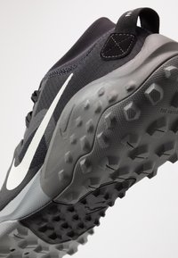 Zwarte en grijze sportschoen met witte Nike swoosh, structuurzool met rubberen noppen en een lusje aan de hiel tegen een witte achtergrond.