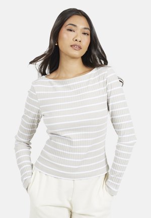 Vrouw met lang donker haar, gekleed in een beige-wit gestreept shirt met lange mouwen en crème-kleurige broek, staand met handen in haar zakken tegen een witte achtergrond.