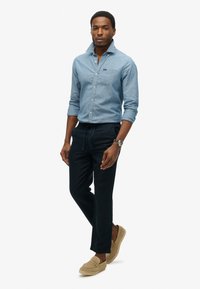 Camicia di denim azzurro chiaro con bottoni e tasca sul petto, abbinata a pantaloni neri scuri con cordoncino e mocassini beige. Texture lisce e design aderente.