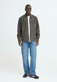 Mann steht in einem grauen Hemd mit Knöpfen über einem weißen T-Shirt, blauen Jeans und schwarzen Turnschuhen mit weißen Streifen.