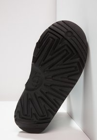 Suela de zapato de goma negra con un patrón de camino en zigzag y el logo "UGG". La superficie tiene un acabado texturizado para mayor agarre y durabilidad.