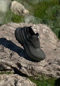 Melnas pārgājienu kurpes ar plūdruma dizainu, teksturētu virsmu, elastīgu potītes atbalstu un izturīgu zoli, novietotas uz akmens virsmas.