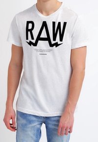 Světle šedé tričko s krátkým rukávem a velkými černými písmeny "RAW" s ostrými akcenty, má pohodlný kulatý výstřih a měkkou texturu látky.