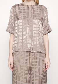 Top beige in satin con un motivo geometrico, maniche corte e scollatura rotonda. Il tessuto ha una texture liscia e una leggera lucentezza.