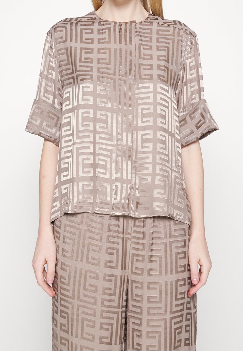 Top beige in satin con un motivo geometrico, maniche corte e scollatura rotonda. Il tessuto ha una texture liscia e una leggera lucentezza.
