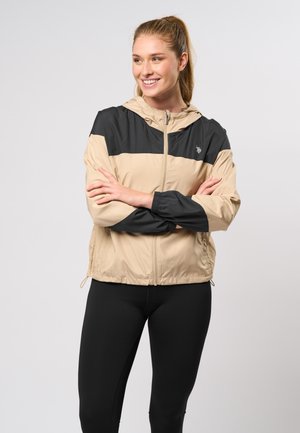 Beige en zwarte windbreaker jack met een capuchon, voorzien van elastische manchetten, een voorrits en een logo op de borst. Glad, lichtgewicht stof.