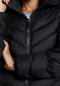 Schwarze Steppjacke mit Reißverschluss vorne, Chevron-Steppmuster und Kapuze. Weiche, gesteppte Textur mit versteckten Taschen.