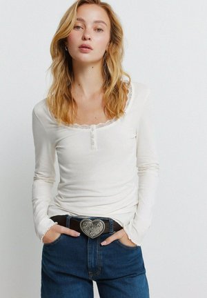 Młoda kobieta o blond włosach, ubrana w biały długi rękaw i niebieskie dżinsy z brązowym paskiem z dużą ozdobną klamrą w kształcie serca.