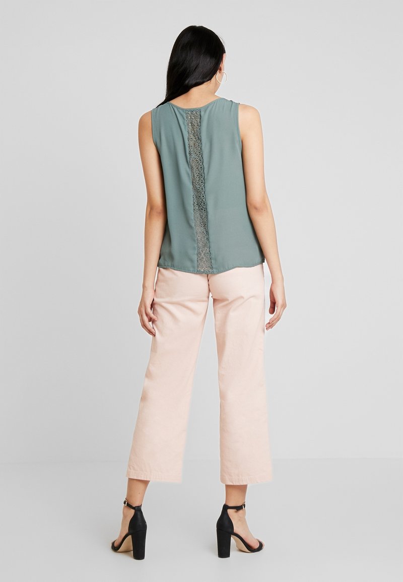Femme portant un haut sans manches turquoise avec un détail en dentelle dans le dos, un pantalon large rose clair et des sandales noires à talons hauts, debout dos à la caméra.