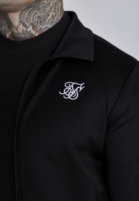 Crna zip-up jakna od glatkog materijala, s visokim ovratnikom i bijelim izvezenim logom na lijevoj strani prsiju. Teksturalna površina i uski kroj.