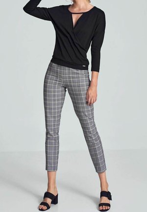 Mujer que lleva una camiseta negra de manga larga con escote en forma de ojo de cerradura, pantalones entallados de cuadros grises y sandalias negras con tacón, de pie contra un fondo claro.