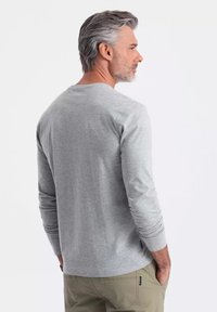 Vaaleanharmaa pitkähihainen puuvillapaita, jossa on pyöreä pääntie ja istuvat hihat, yhdistettynä vaaleisiin housuihin. Takakulman näkymä vaatteesta.