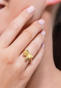 Goldring mit floralem Design und strukturierten Blütenblättern, auf einer Hand, die sanft das Gesicht berührt. Glatte Haut und heller Nagellack sind sichtbar.