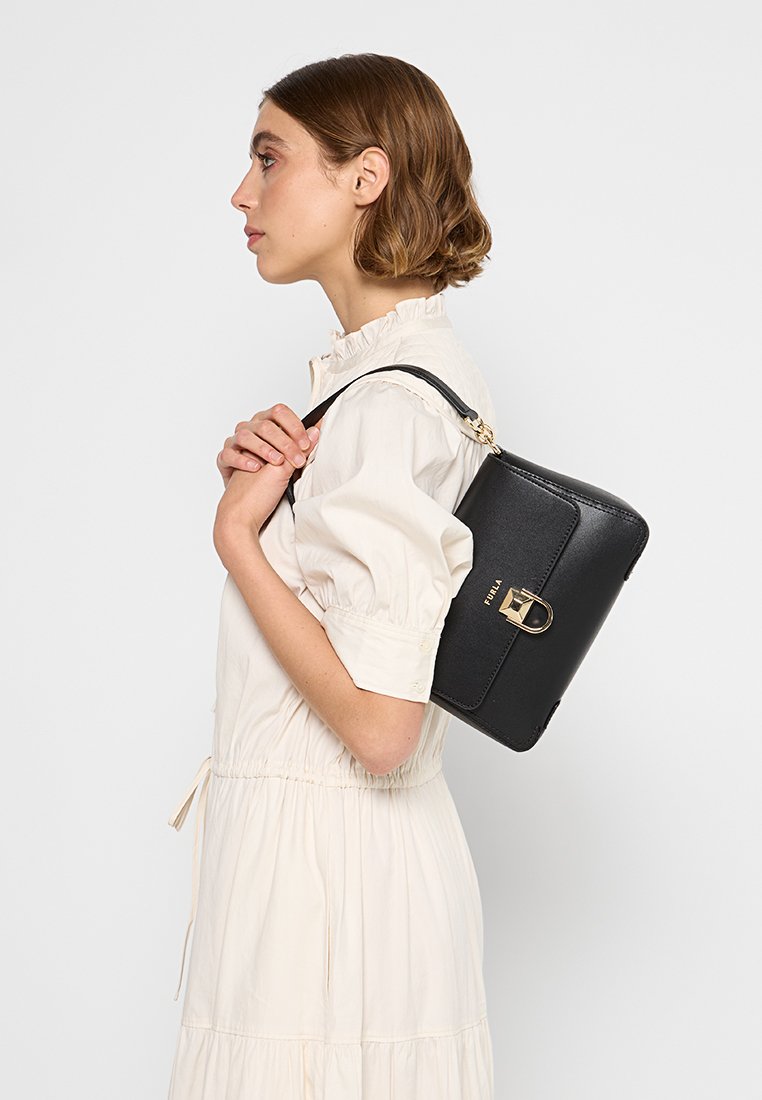 Sac à main rectangulaire noir avec une finition texturée, doté d'un fermoir doré et d'une courte anse, porté sur l'épaule contre une robe claire.