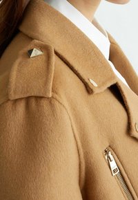 Kamelowy wełniany płaszcz z ozdobnymi złotymi elementami, zapinanymi na guziki patkami na ramionach i ukośną kieszenią na zamek. Miękka faktura i dopasowany krój.