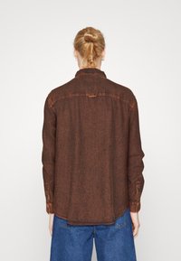 Brun denimskjorta med långa ärmar, knappmanschetter och krage. Har en texturerad yta och subtila stygn på baksidan.