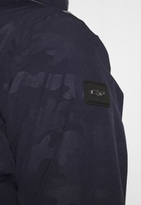 Marinejakke med et tekstureret stof og et subtilt camouflage-mønster. Inkluderer en sort lap med et logo på skulderen.