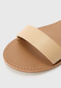 Brun slide sandal med en bred, glat beige rem og en simpel brun sål med synlige søm langs kanten.