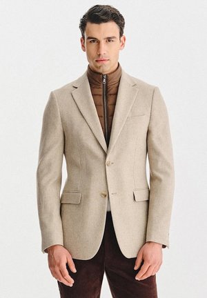 Beżowa sztruksowa marynarka z klapami z wycięciem, dwoma guzikami i przednimi kieszeniami, noszona na brązowej puchowej kurtce z wysokim kołnierzem i zamkiem błyskawicznym.