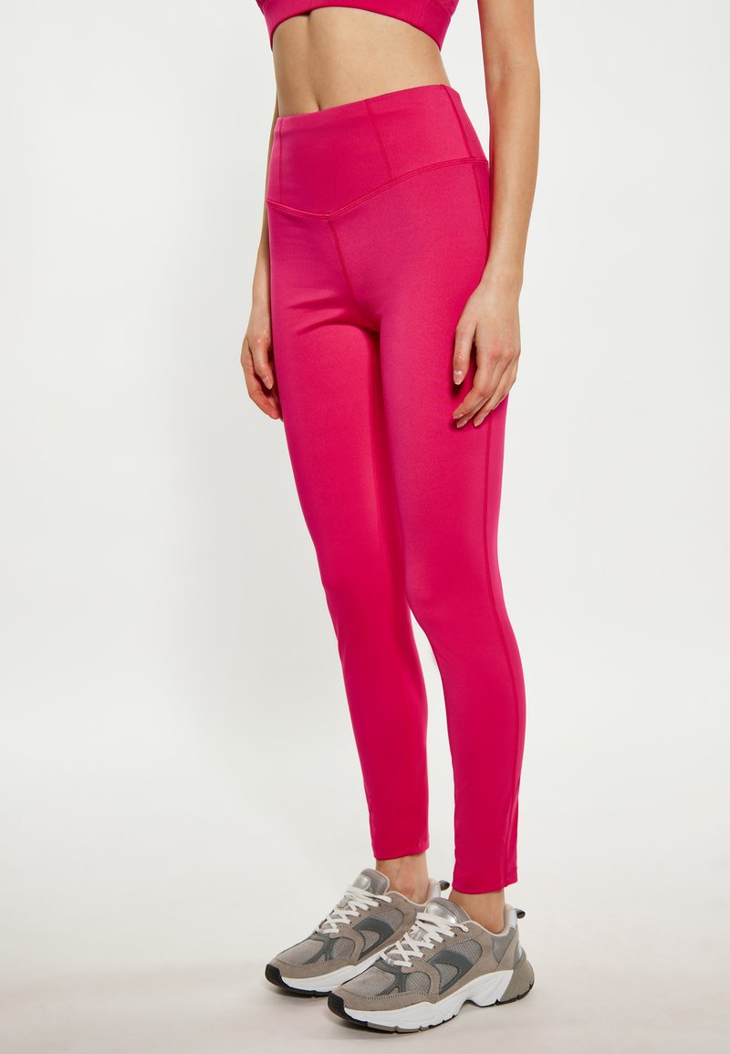 Leggings aderenti di materiale rosa brillante con vita alta, caratterizzati da cuciture piatte. Abbinati a sneaker grigie con accenti bianchi.