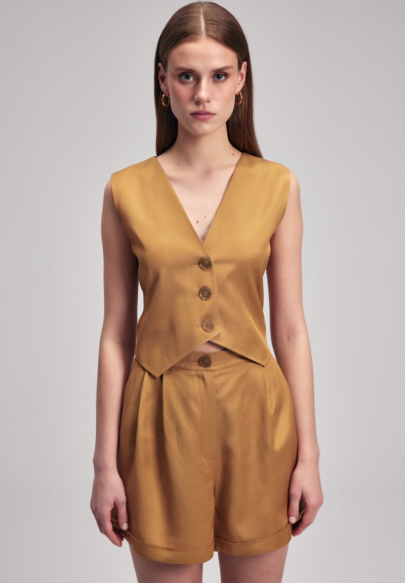 Chaleco sin mangas mostaza con cuello en V, que cuenta con tres botones marrones. Combinado con pantalones cortos de cintura alta a juego, ambos de textura suave.