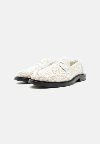 Paire de mocassins blancs texturés avec semelles noires, dotés d'une bride lisse sur le dessus, sur un fond clair uni.