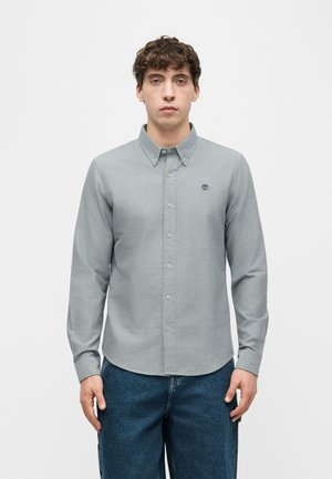 Jeune homme portant une chemise à manches longues gris clair boutonnée avec col et un jean bleu, debout devant un fond blanc uni.