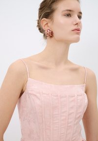 Top corsé rosa con textura floral y plisados verticales; modelo lleva pendientes redondos con gemas rosas y herrajes dorados.