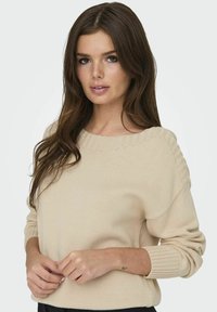 Beige kötött pulóver széles nyakkivágással, bordás mandzsettákkal és diszkrét textúrával. A modell a kezeit a hasa előtt tartja, bemutatva a laza szabást.