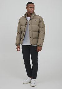 Beige puffer jacket with a high collar, featuring a button front and side pockets, paired with a white shirt and black pants.