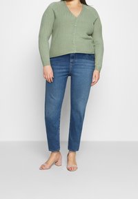 Vihreä ribbineulottu neuletakki V-kaula-aukkolla ja nappikiinnityksellä, yhdistetty sinisiin farkkuihin ja vaaleanpunaisiin korkosandaaleihin.