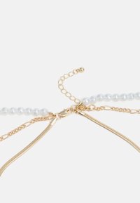 Correntes em tom dourado com elos texturizados e um fecho, combinadas com uma fila de contas redondas azul-claro. Vários elementos entrelaçam-se elegantemente.