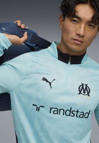 Haut de sport bleu clair avec une demi-fermeture éclair, présentant un motif texturé, un logo Puma noir, un emblème de Marseille et un marquage "randstad".
