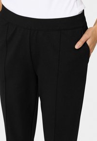 Black trousers with a smooth texture, featuring a wide waistband, subtle pleats, and side pockets for practical use.