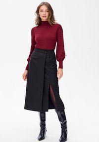 Bordeaux aansluitende trui met hoge hals, gecombineerd met een zwarte A-lijn midi rok met een voorin gespleten voorkant en elegante zwarte hoog aangesloten laarzen.