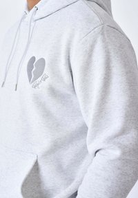 Sudadera gris texturizada con un logotipo de corazón roto en gris oscuro y firma. Cuenta con cordones ajustables y un bolsillo en la parte frontal.