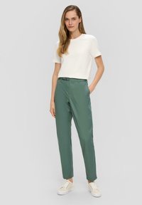 Leichtes, weißes Baumwoll-T-Shirt mit kurzen Ärmeln, kombiniert mit grünen schmal geschnittenen Hosen und beigen Sneakern. Flaches Vorderdesign, schlichte Eleganz.