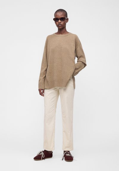 Personne portant un pull marron clair oversize, un pantalon crème, des chaussures rouge foncé avec des lacets blancs et des lunettes de soleil foncées, debout devant un fond uni.
