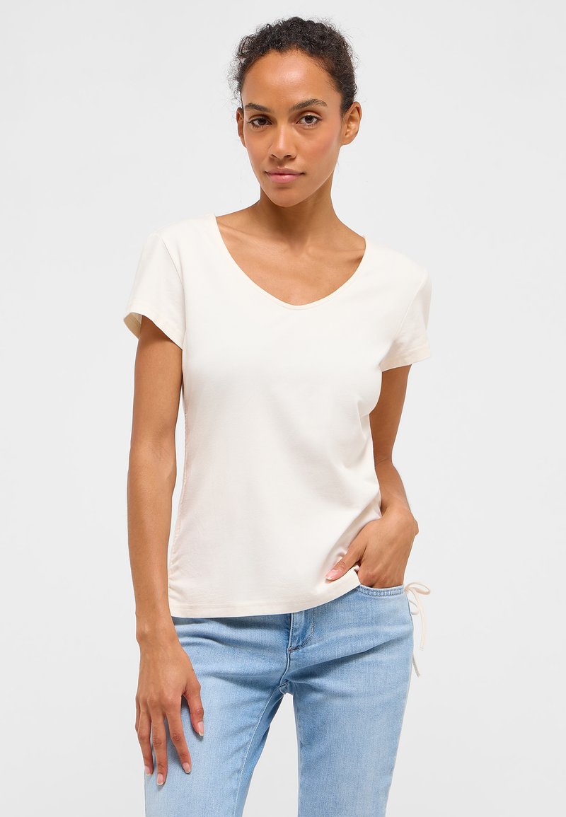 Žena s kučeravými vlasmi v bielej tričku s krátkym rukávom a véčkovým výstrihom a svetlomodrými džínsami, stojaca s jednou rukou vo vrecku.