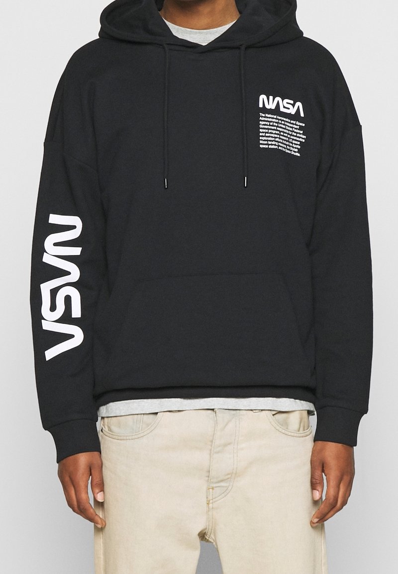 Sudadera negra con capucha con el logo y texto blanco de la NASA en el pecho, texto grande blanco de la NASA en la manga izquierda, combinada con pantalones beige claro.