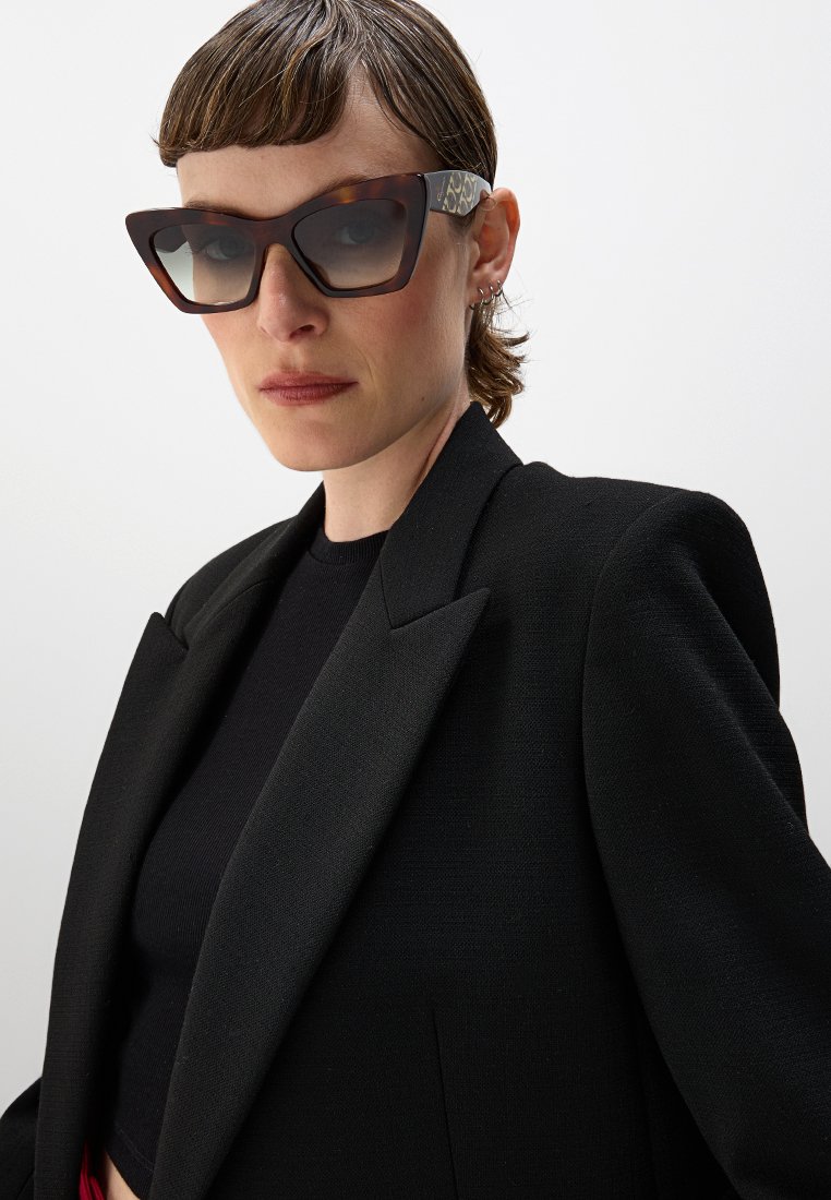 Gafas de sol carey con montura angular y diseño ancho; cuentan con patillas estampadas y lentes con suave degradado. Blazer y top negros.