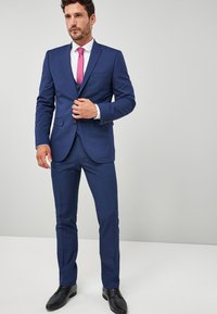 Marineblauer Anzug mit strukturiertem Blazer und schmal geschnittenen Hosen. Besitzt eine kontrastierende rosa Krawatte, ein weißes Hemd und schwarze Dressschuhe.