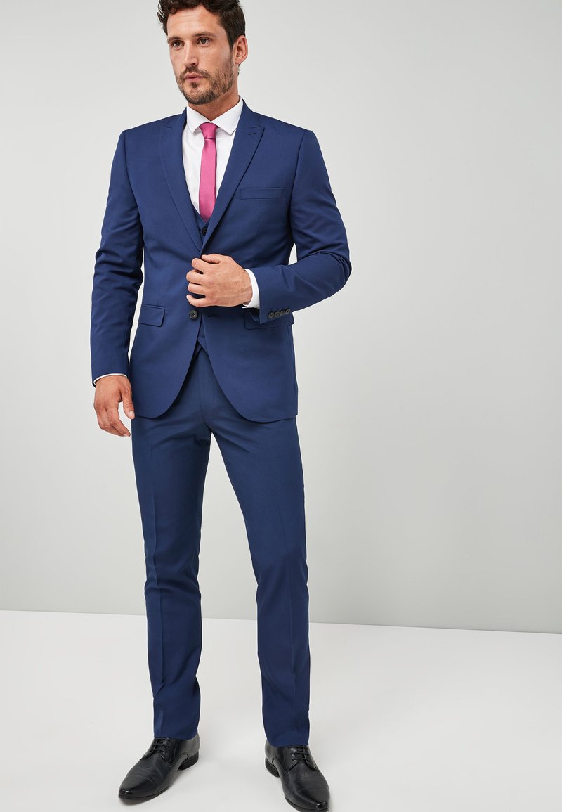 Marineblauer Anzug mit strukturiertem Blazer und schmal geschnittenen Hosen. Besitzt eine kontrastierende rosa Krawatte, ein weißes Hemd und schwarze Dressschuhe.