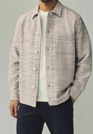 Man wearing textured beige button-up jacket over white shirt with dark pants, standing with one hand in pocket against neutral background.