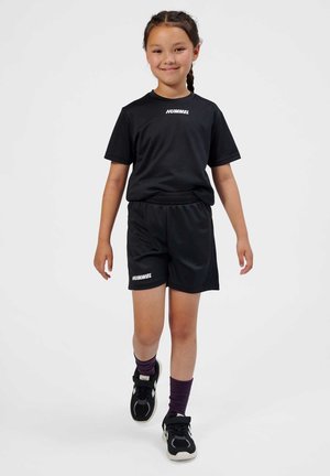 Ragazza che cammina in avanti indossando abbigliamento sportivo nero Hummel, scarpe da ginnastica nere e calzini viola, sorridendo con i capelli intrecciati su sfondo bianco.