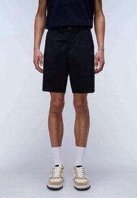 Μαύρες βερμούδες σε χαλαρή γραμμή, με κορδόνι στη μέση. Φορεμένες με μπεζ και λευκά αθλητικά παπούτσια, μαζί με λευκές αθλητικές κάλτσες.