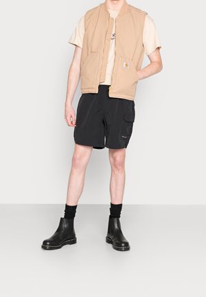 Gilet en tissu beige porté sur un t-shirt beige clair, associé à un short cargo noir et des bottines noires à cheville avec des côtés élastiques et des détails de couture.