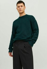 Pull en maille vert foncé avec un col rond et des poignets côtelés, associé à un pantalon noir. Texture douce, coupe décontractée et couleur unie.