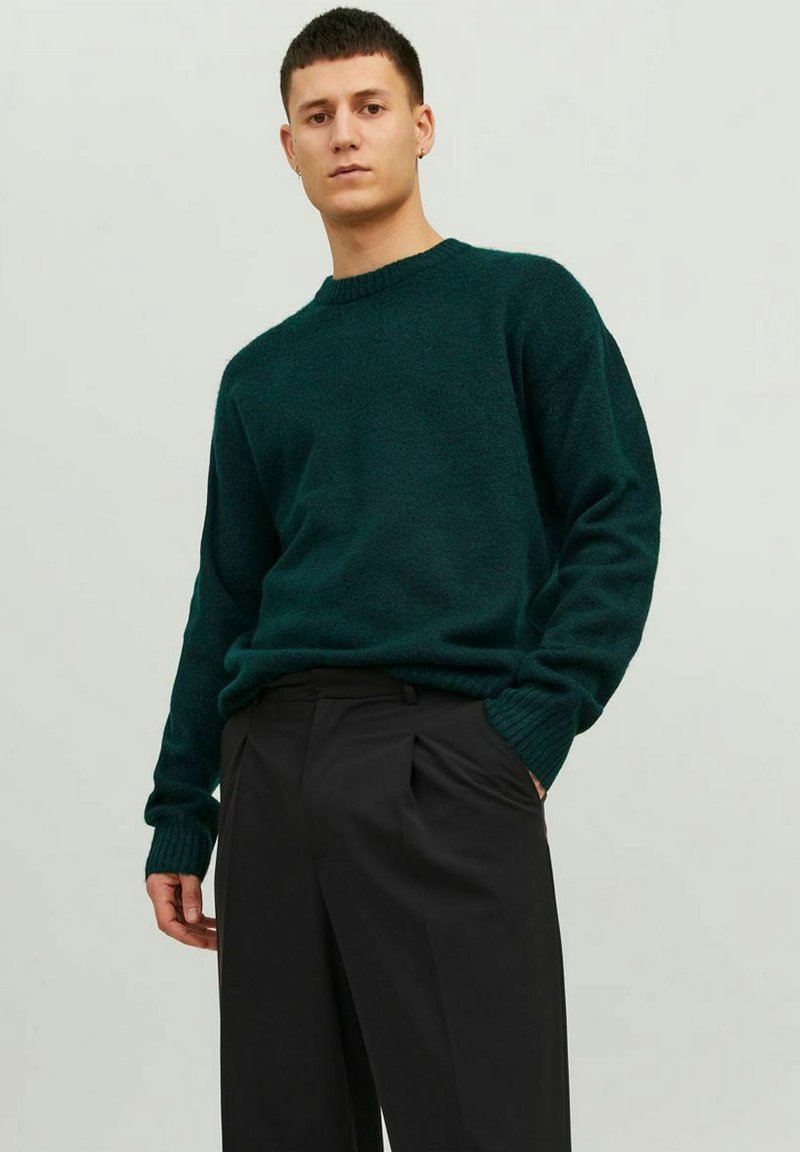 Pull en maille vert foncé avec un col rond et des poignets côtelés, associé à un pantalon noir. Texture douce, coupe décontractée et couleur unie.